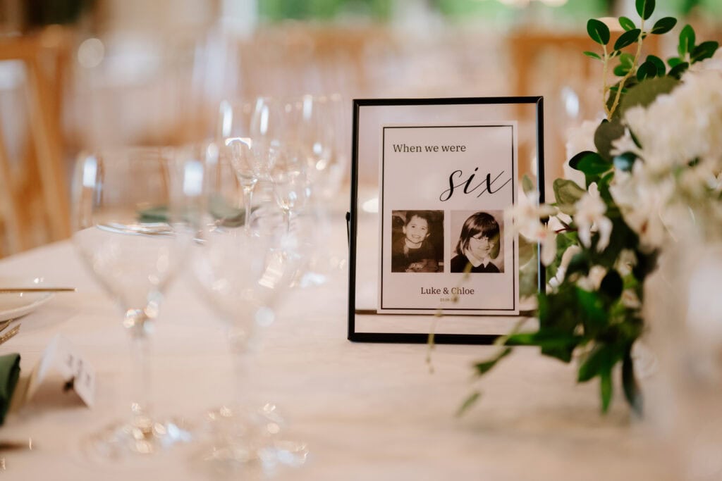 A wedding table at The Old Kent Barn is set with elegant dinnerware and glassware. A framed photo titled "When we were six" shows two children. Nearby, a lush green and white floral centerpiece enhances the charming decor. Image by Pearce Wedding Photography.