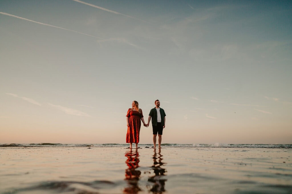 A couple holds hands during their engagement photography session in Kent, walking on a calm, reflective beach at sunset. The sky is clear with a few wispy clouds, and the ocean waves gently lap at their feet, casting reflections on the wet sand. Image by Pearce Wedding Photography.