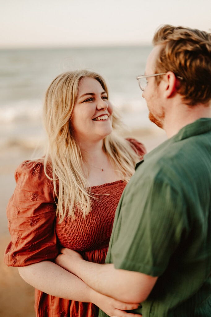 A couple stands on a beach, gazing at each other with smiles during their pre-wedding photoshoot. The woman wears a rust-colored dress, and the man wears a green shirt. The ocean waves and sky form the perfect romantic backdrop for this special moment. Image by Pearce Wedding Photography.