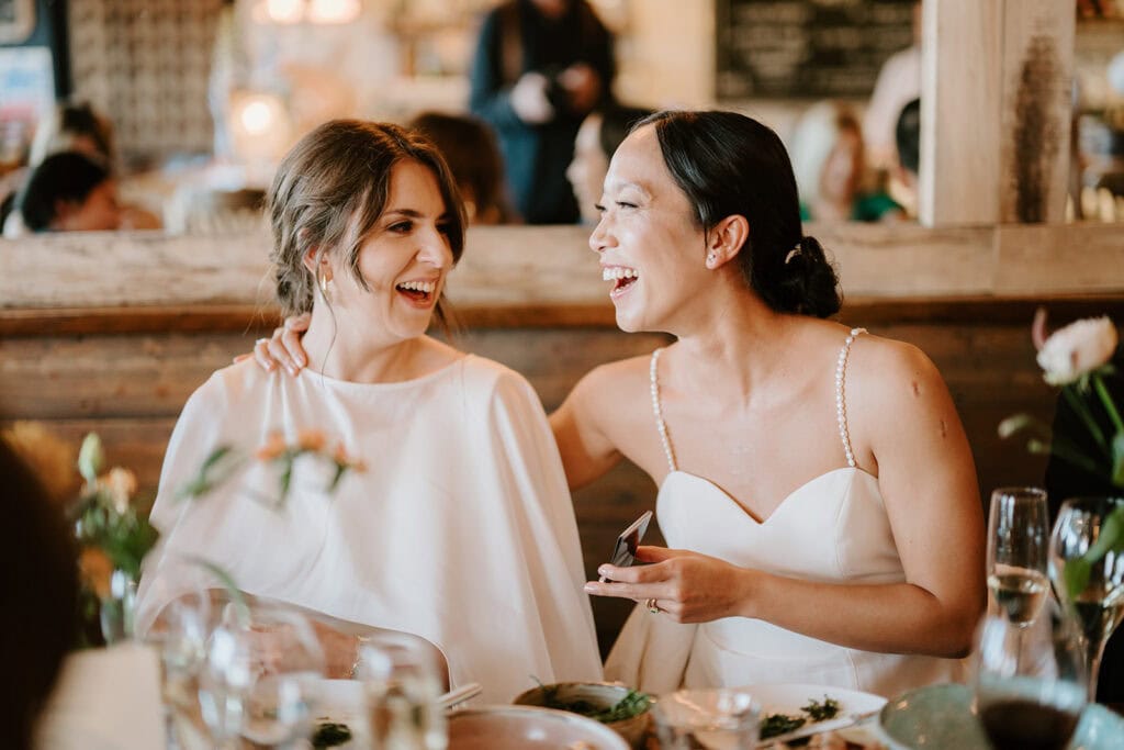 Two women in elegant white dresses smile and sit closely together at a dining table during a wedding celebration. They appear happy and are engaged in conversation, surrounded by plates and glasses, adding warmth to the joyous Broadstairs gathering. Image by Pearce Wedding Photography.