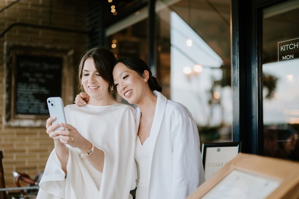 Two women, dressed in elegant white outfits, smile and take a selfie outside a building in Broadstairs. One has shoulder-length brown hair, while the other sports long dark hair tied back. They seem to be relishing a casual moment during what seems to be a wedding celebration. Image by Pearce Wedding Photography.