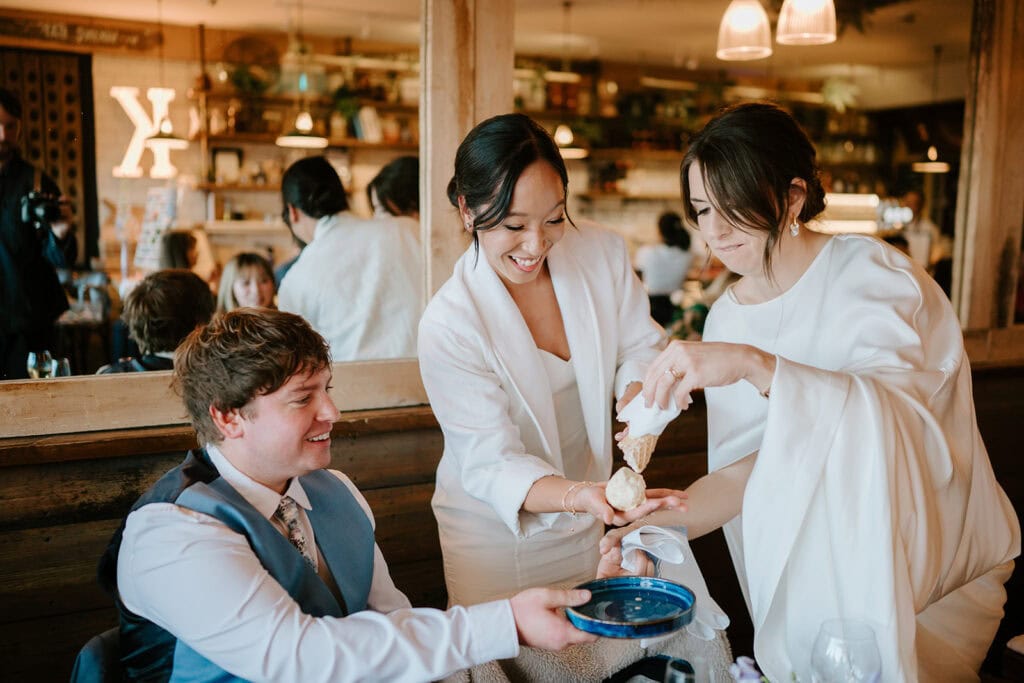 Two women in white outfits happily serve food to a seated man in a blue vest at a lively Broadstairs restaurant. Behind them, shelves display various bottles and decorations. The atmosphere is warm and welcoming, reminiscent of a joyful wedding celebration. Image by Pearce Wedding Photography.