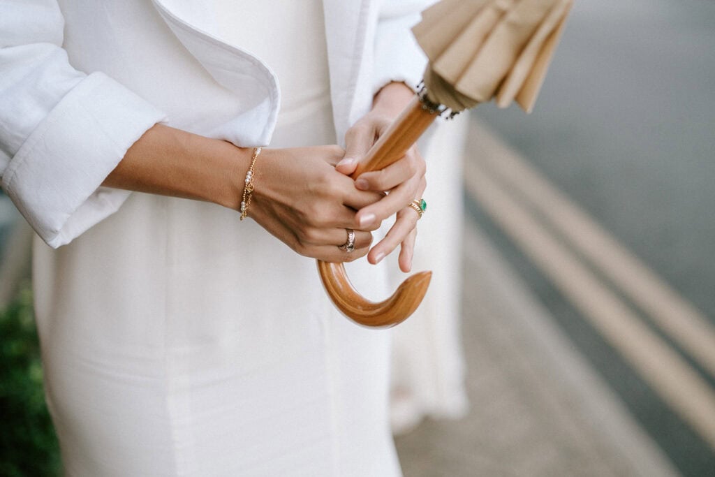 A person in a white outfit, possibly on their way to a wedding, holds a closed beige umbrella with a wooden handle. Adorned with bracelets and rings, they stand on a sidewalk near the double yellow lines of Broadstairs' charming streets. Image by Pearce Wedding Photography.