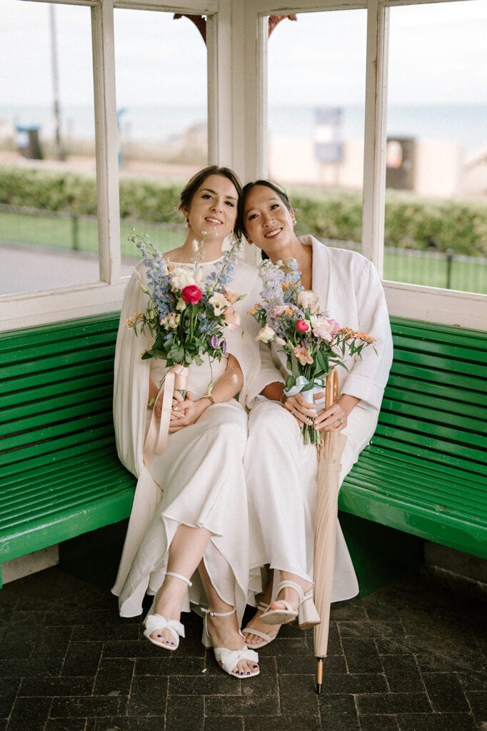 Two women in white wedding dresses, holding bouquets of flowers, sit together on a green bench inside a Broadstairs gazebo. One holds a closed umbrella. They both smile warmly at the camera. Image by Pearce Wedding Photography.