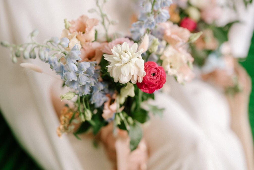 A close-up of a wedding floral arrangement featuring delicate blooms. The bouquet includes a white dahlia, a vibrant pink ranunculus, and soft blue delphiniums, all nestled among light pink and green accents against a blurred background reminiscent of Broadstairs' coastal charm. Image by Pearce Wedding Photography.