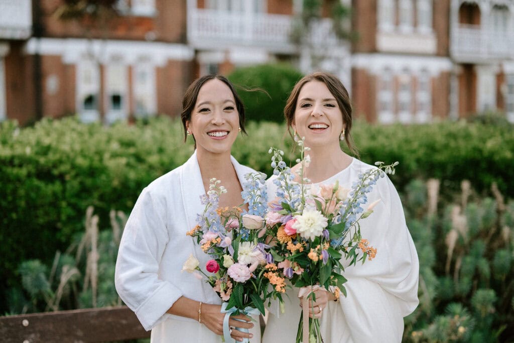 Two women are standing outdoors in front of a garden, wearing white outfits and radiant wedding smiles. They hold bouquets of colorful flowers, with a charming Broadstairs building providing a picturesque backdrop. Image by Pearce Wedding Photography.
