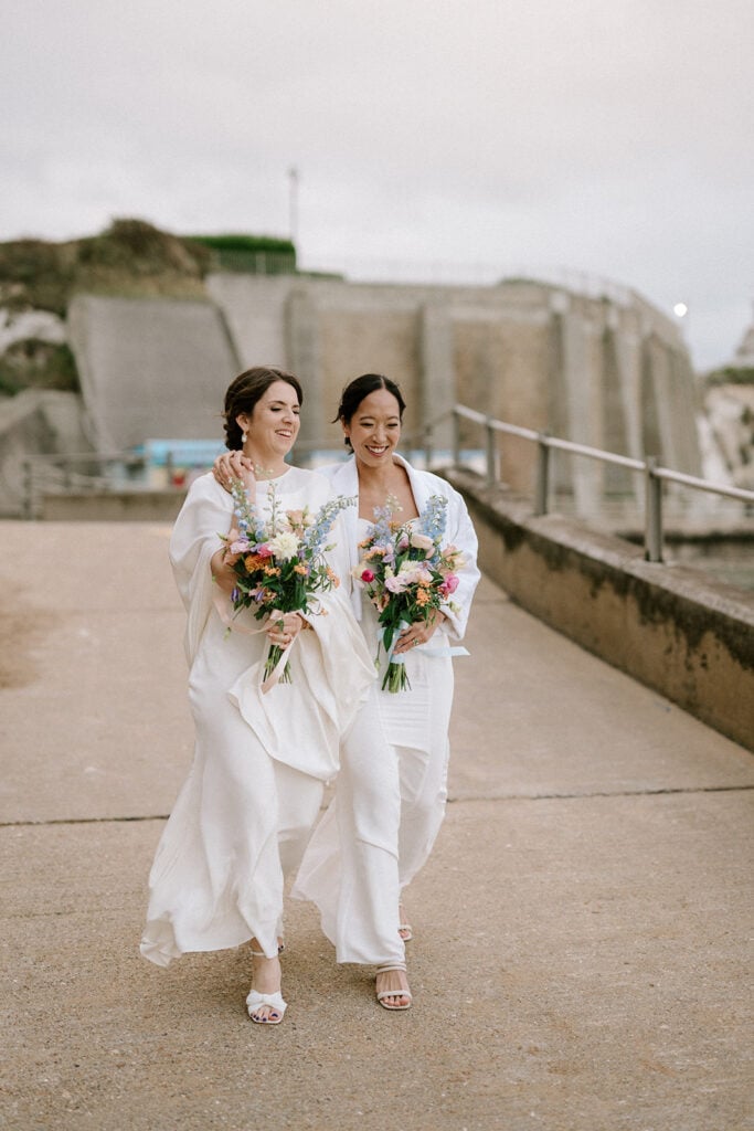 Two women in white dresses walk arm-in-arm, smiling and holding colorful bouquets. They are near a coastal area in Broadstairs, with concrete structures in the background under a cloudy wedding sky. Image by Pearce Wedding Photography.