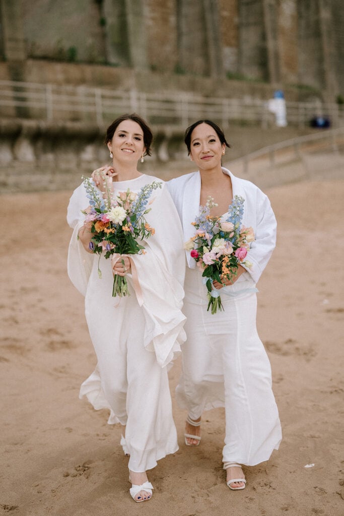 Two individuals in white outfits walk along Broadstairs beach, each holding a colorful bouquet of flowers. They smile joyfully, the ocean and sand setting the perfect backdrop. With one having an arm around the other, it feels like a serene wedding moment by the sea. Image by Pearce Wedding Photography.