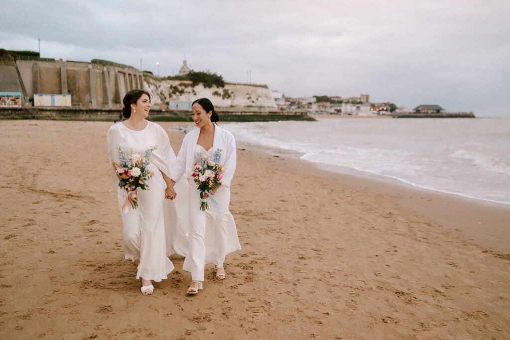 Two people in white outfits walk hand in hand on a sandy Broadstairs beach, each holding a vibrant bouquet of flowers. The backdrop features coastal buildings and an overcast sky, enhancing the serene and intimate wedding atmosphere. Image by Pearce Wedding Photography.