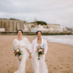 Two women in white outfits walk hand in hand on a beach, each holding a bouquet of colorful flowers. Cliffs and a lighthouse are visible in the background, under a cloudy sky captured by a talented wedding photographer in Kent. They are both smiling and barefoot on the sand. Image by Pearce Wedding Photography.