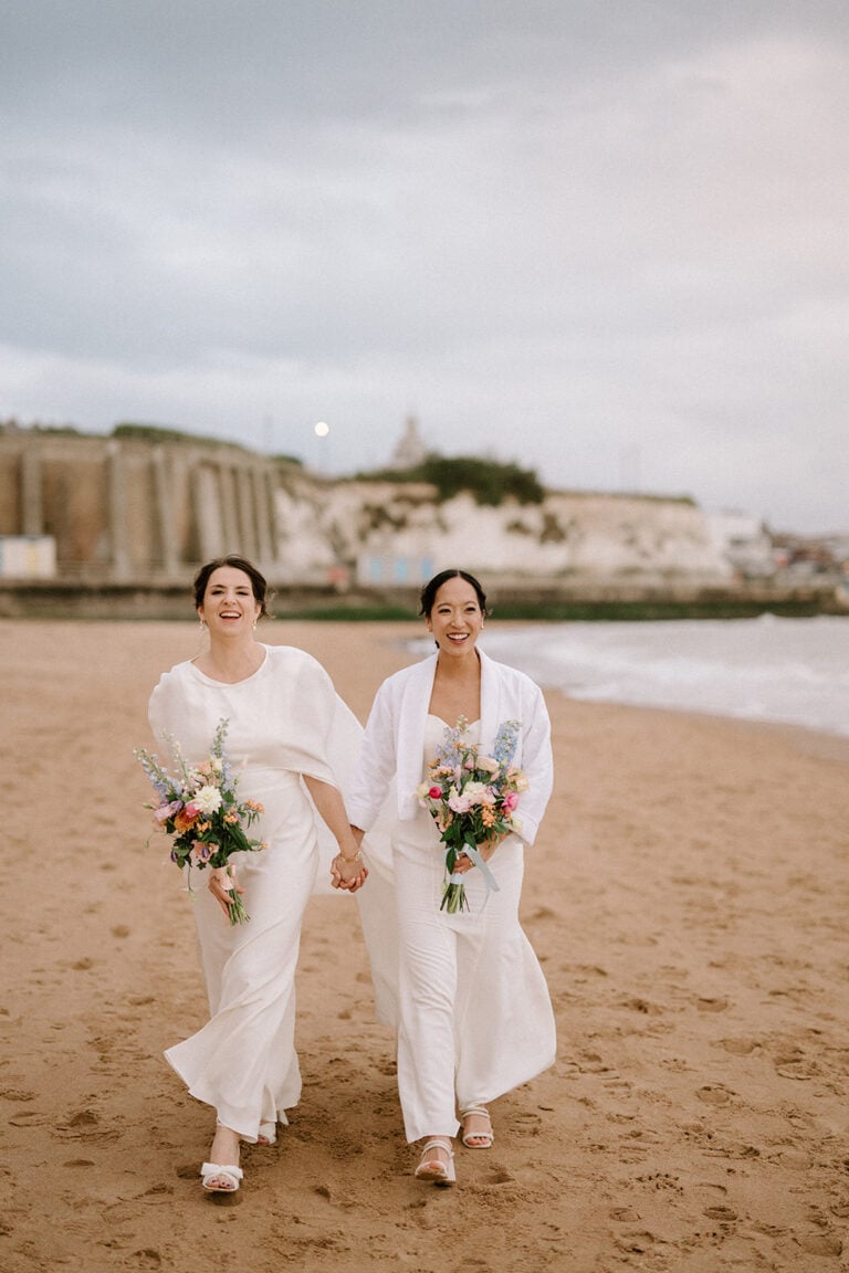 Two women in white outfits walk hand in hand on a beach, each holding a bouquet of colorful flowers. Cliffs and a lighthouse are visible in the background, under a cloudy sky captured by a talented wedding photographer in Kent. They are both smiling and barefoot on the sand. Image by Pearce Wedding Photography.