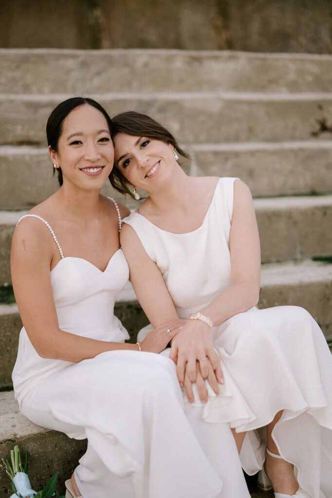 Two women in elegant white dresses sit closely on stone steps, smiling warmly, as if capturing a candid moment at a Broadstairs wedding. One leans affectionately on the other's shoulder. The outdoor setting adds a natural and relaxed ambiance to the scene. Image by Pearce Wedding Photography.