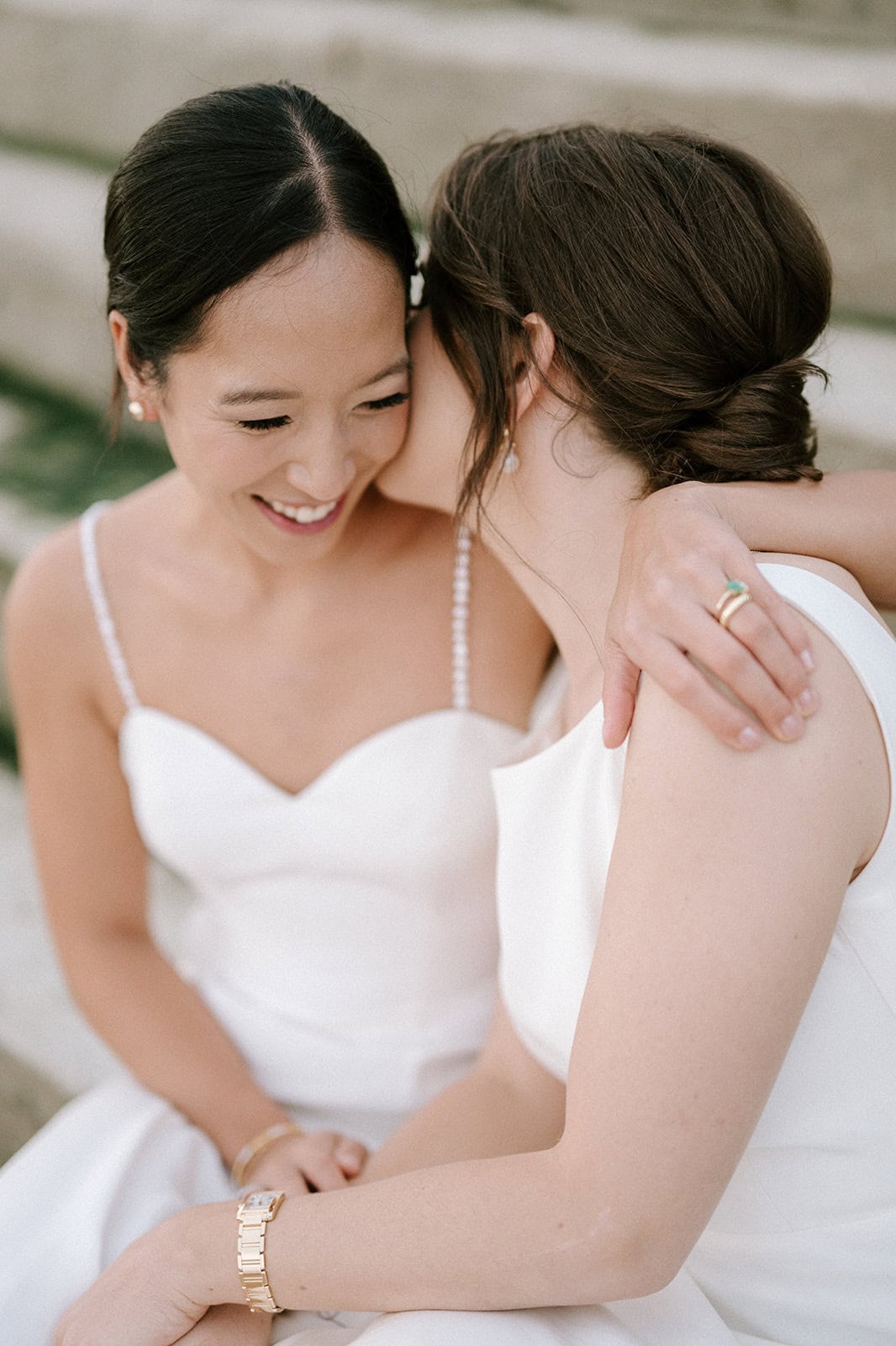 On the stone steps of Broadstairs, two women in white dresses share a joyful moment. One whispers into the other's ear, both smiling as their arms gently entwine. It's as if they are savoring a secret at an intimate wedding celebration. Image by Pearce Wedding Photography.