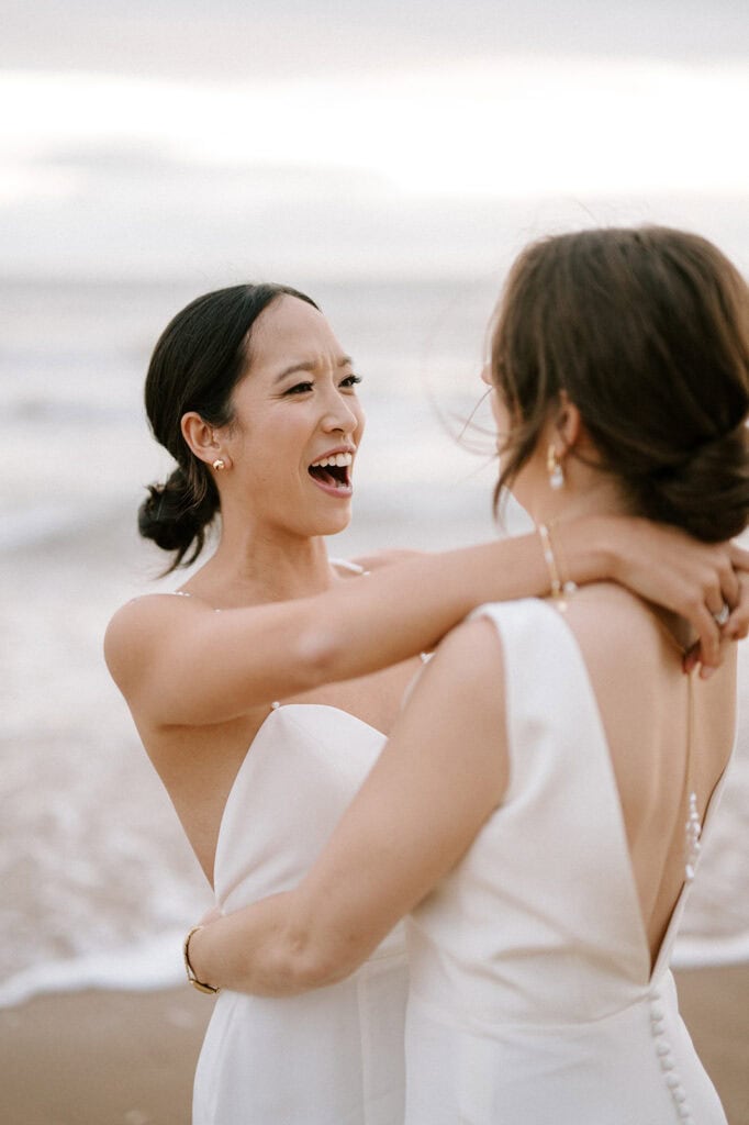 Two women in white wedding dresses embrace joyfully on a Broadstairs beach, with the ocean and sky forming a serene backdrop. Smiling, they celebrate their special wedding moment together. Image by Pearce Wedding Photography.