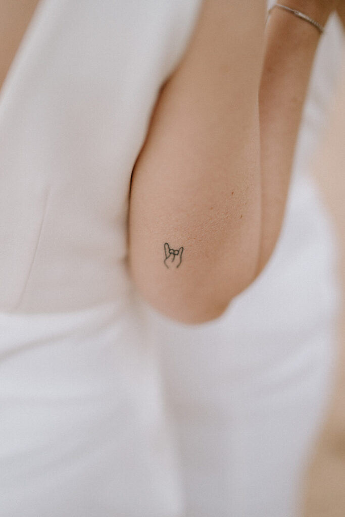 In a white sleeveless outfit at a Broadstairs event, a close-up captures a person's arm adorned with a small tattoo—a hand making the rock 'n' roll gesture on the inner side. Image by Pearce Wedding Photography.