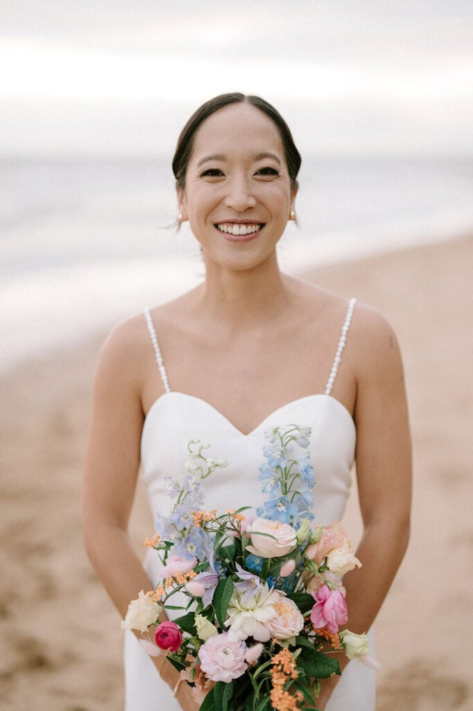 A woman in a white dress stands on the beach at Broadstairs, smiling while holding a colorful bouquet of flowers. The ocean and sandy shore are blurred in the background, capturing the perfect wedding moment. Image by Pearce Wedding Photography.