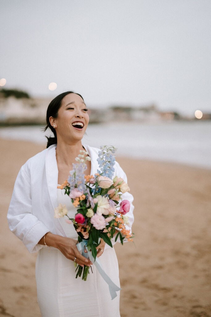 A joyful woman in a white dress, possibly a bride, holds a colorful bouquet while standing on the sandy beach of Broadstairs. The blurred ocean and distant buildings form the perfect backdrop against the soft evening sky for this serene wedding scene. Image by Pearce Wedding Photography.
