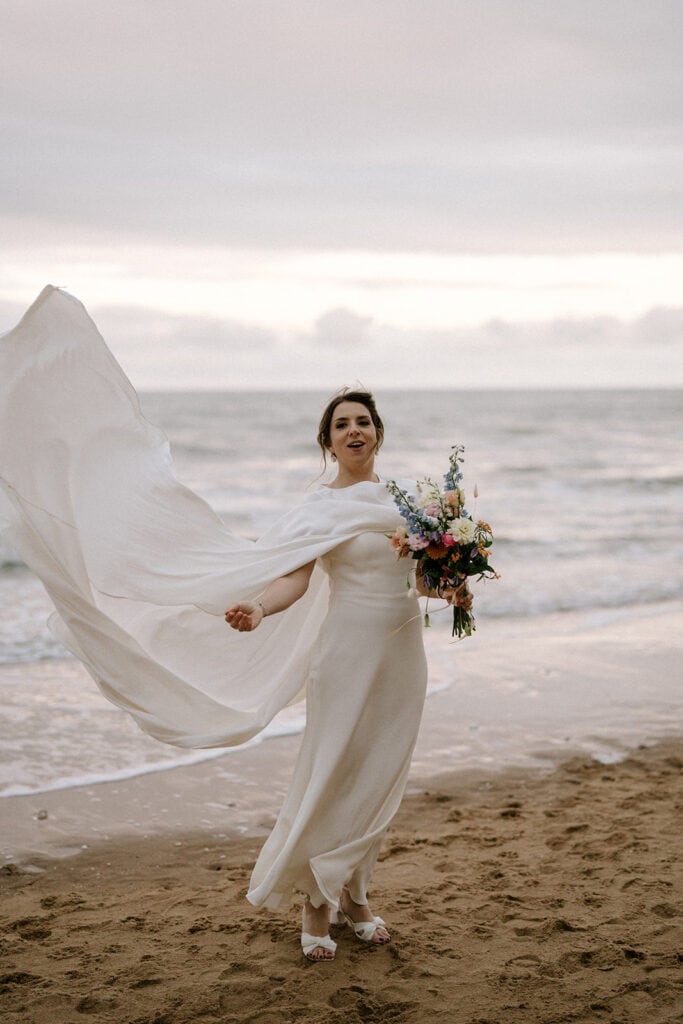 A person in a flowing white dress stands joyfully on the sandy beach at Broadstairs, holding a colorful bouquet. The ocean waves and a cloudy sky provide a serene and picturesque backdrop, creating the perfect setting for an enchanting wedding moment. Image by Pearce Wedding Photography.