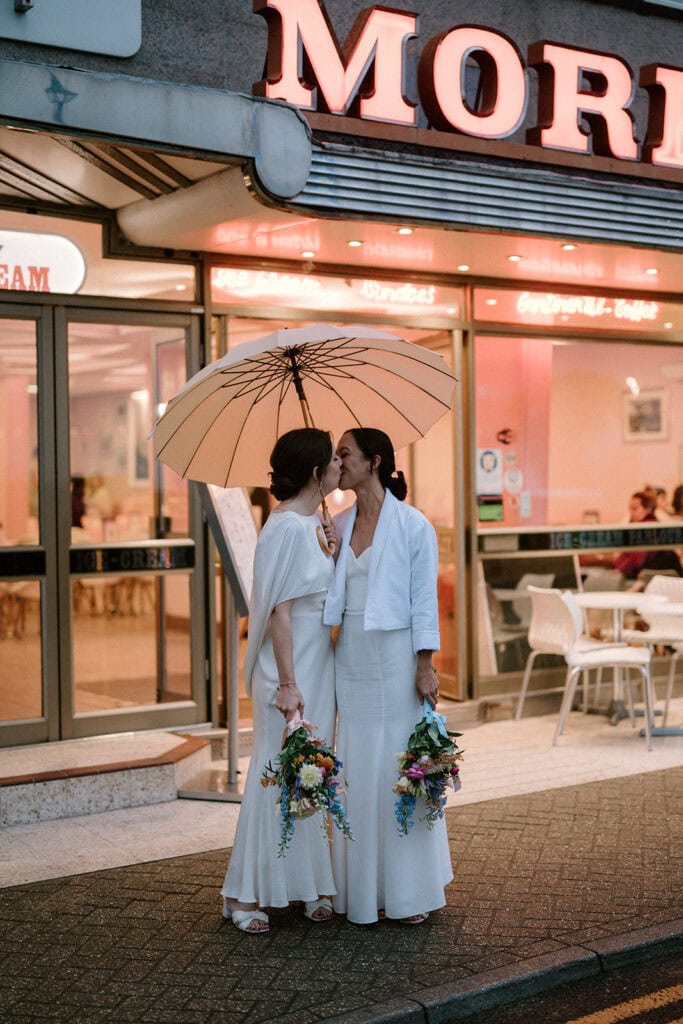 Two women in white wedding outfits share a kiss under a beige umbrella outside a retro-style ice cream shop in Broadstairs. Each holds vibrant flower bouquets, standing on the sidewalk with a neon sign glowing above them, capturing the essence of their love and celebration. Image by Pearce Wedding Photography.