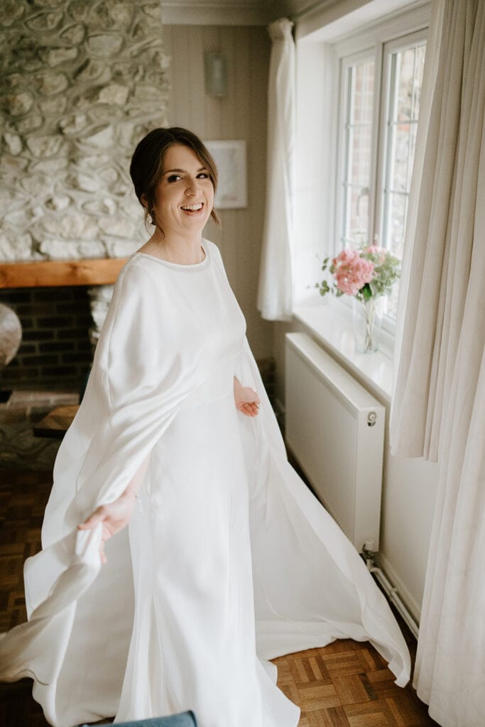 A woman in a flowing white dress smiles while standing near a window with curtains, as if preparing for her Broadstairs wedding. A bouquet of pink flowers rests on the sill, surrounded by a stone wall and wooden floor. Natural light brightens the space. Image by Pearce Wedding Photography.