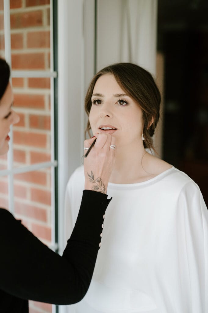 A bride in a stunning white outfit has her makeup done by an artist, their hands carefully applying lipstick. Indoors, against a brick wall with a window, the scene captures the elegance of a Broadstairs wedding preparation. Image by Pearce Wedding Photography.