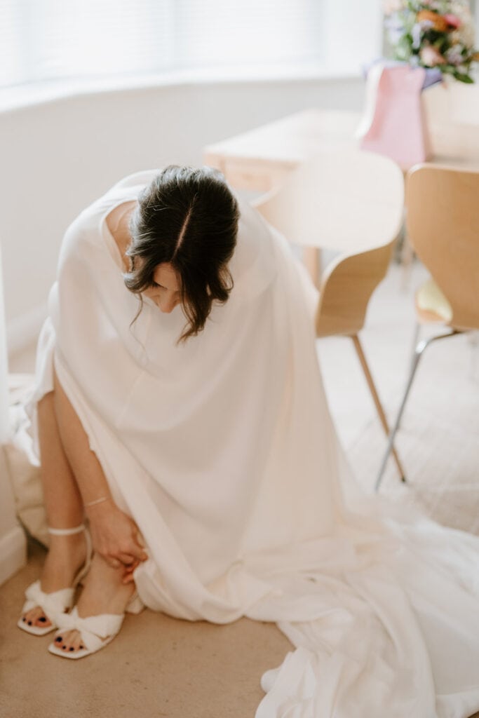 A bride in a white dress sits in a softly lit room, adjusting her shoe for the perfect wedding day. Her hair is styled in an elegant updo, surrounded by subtle, modern furniture reminiscent of Broadstairs charm. A vibrant bouquet graces the background on a table. Image by Pearce Wedding Photography.
