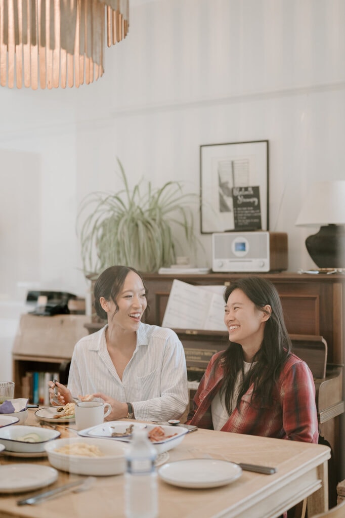 Two people sit at a dining table in Broadstairs, enjoying a meal together. Both are smiling, and a piano with sheet music graces the background. Natural light fills the room, evoking the warm and inviting atmosphere of an intimate wedding celebration. Image by Pearce Wedding Photography.