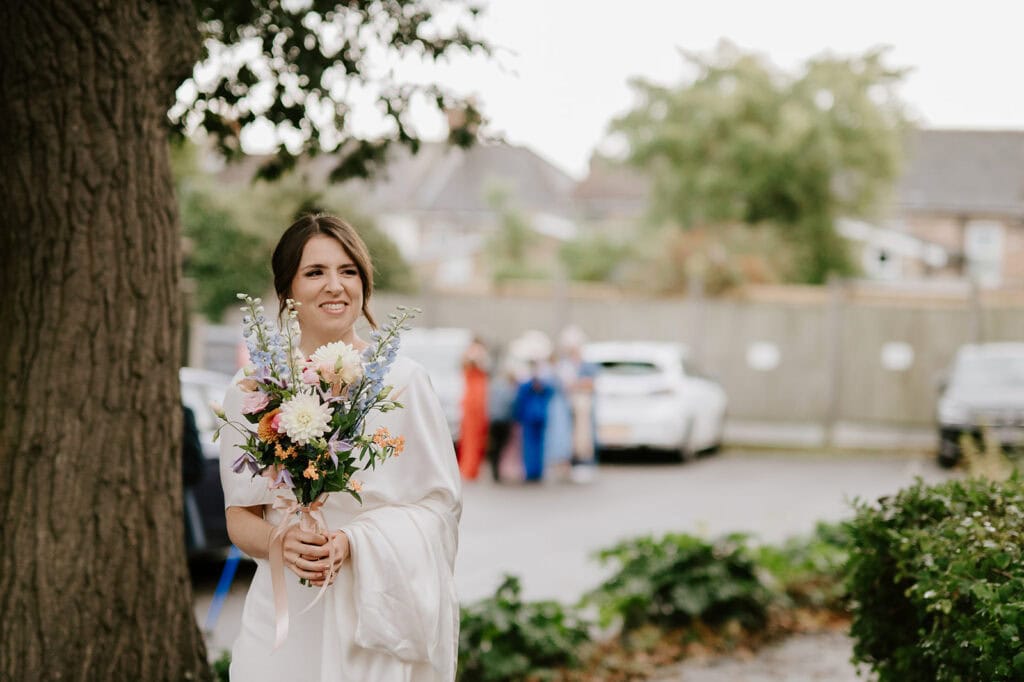 A bride in a white dress, holding a bouquet of colorful flowers, stands outdoors near a tree at her Broadstairs wedding. Blurred in the background are a few people and parked cars on a paved area. Image by Pearce Wedding Photography.