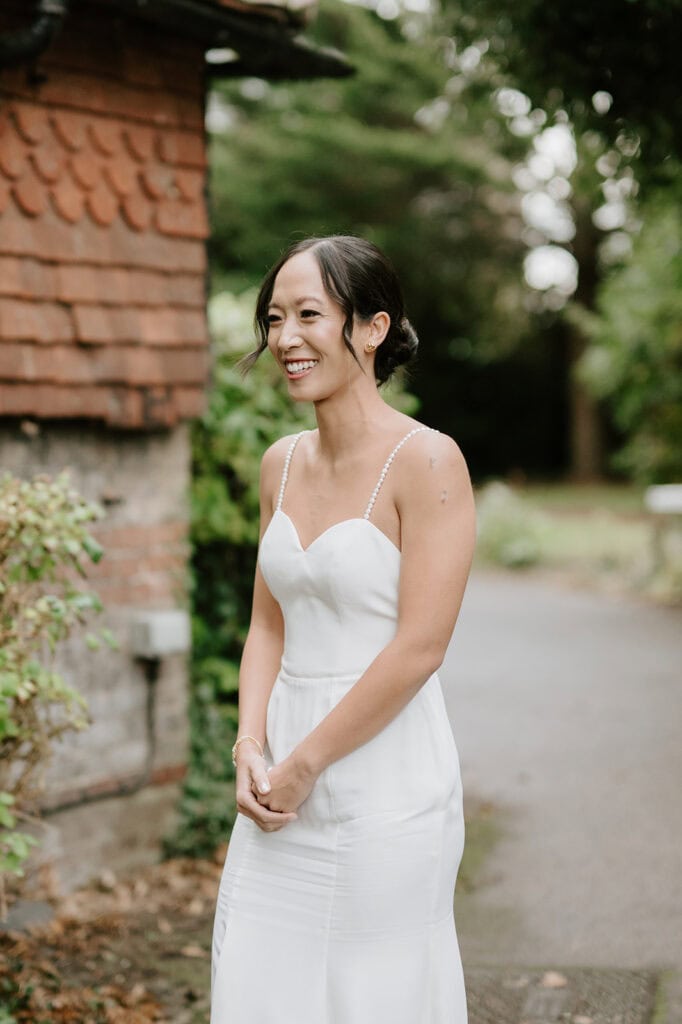 A woman in a white wedding dress stands outdoors, smiling beside a brick building. She has dark hair styled in an updo and is surrounded by Broadstairs greenery. The scene is bright and cheerful. Image by Pearce Wedding Photography.