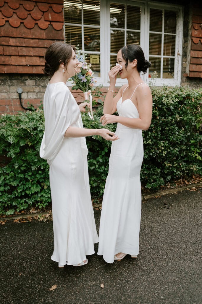 Two women in white dresses stand outdoors, smiling and chatting like two joyful souls at a wedding. One holds a bouquet of flowers, the scene unfolding charmingly in front of a brick building adorned with green bushes and a window, reminiscent of picturesque Broadstairs. Image by Pearce Wedding Photography.
