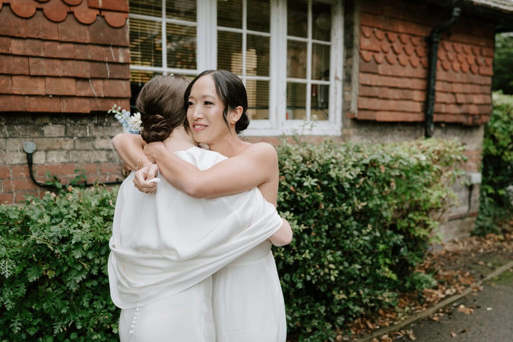 Two people in white dresses embrace in front of a brick building adorned with greenery, capturing a tender wedding moment. The person facing the camera has flowers in their hair, while the other gently places a hand on their shoulder, evoking joy reminiscent of a Broadstairs love story. Image by Pearce Wedding Photography.