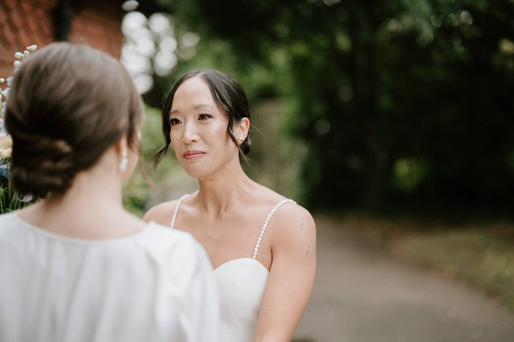 In the lush greenery of Broadstairs, two women in wedding dresses share an intimate moment. One faces away from the camera, while the other smiles gently at her, basking in the soft, natural light. Image by Pearce Wedding Photography.