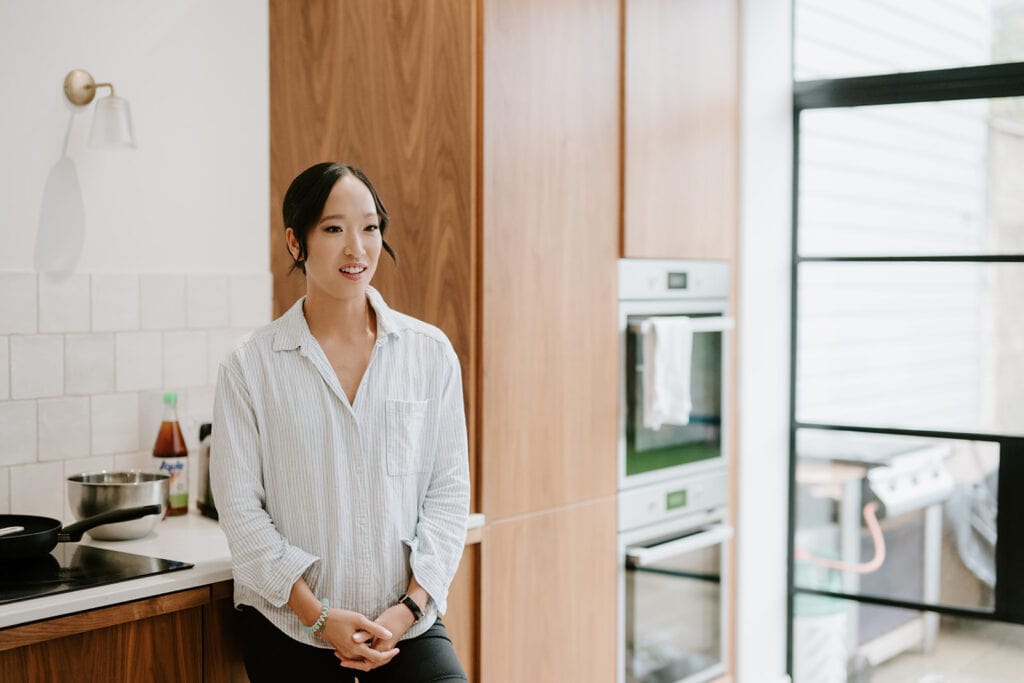 A person with long dark hair, wearing a light striped shirt, leans casually against a wooden kitchen counter. The kitchen, reminiscent of a cozy Broadstairs venue, boasts modern appliances, abundant natural lighting through a large window, and a warm, inviting atmosphere. Image by Pearce Wedding Photography.