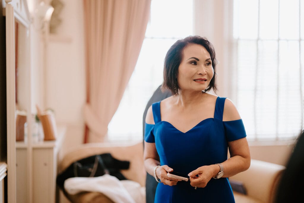 A woman in a blue off-shoulder dress stands indoors at a Broadstairs venue, holding a phone. The room is softly lit, with elegant curtains and a couch in the background. She appears to be smiling slightly, possibly capturing memories of a wedding celebration. Image by Pearce Wedding Photography.