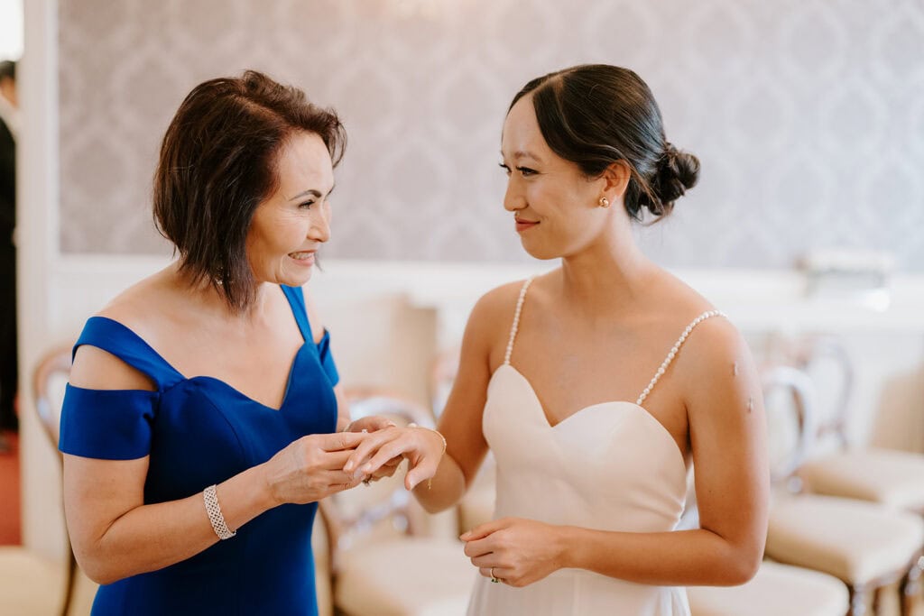 A bride in a white dress shows her ring to a woman in a blue dress. They are smiling and standing in an elegant room adorned with exquisite wallpaper and chairs—capturing the joyous spirit of their Broadstairs wedding. Image by Pearce Wedding Photography.