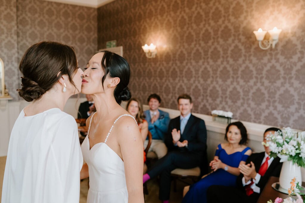 In a beautifully decorated room in Broadstairs, two brides in white gowns share a kiss, surrounded by smiling guests. The wallpaper's intricate pattern complements the elegant floral arrangement on the right, creating a perfect wedding scene. Image by Pearce Wedding Photography.