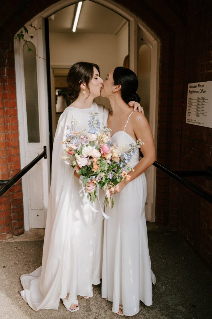 Two brides in white dresses kiss and embrace outside a building in Broadstairs. One holds a colorful bouquet of flowers. The wedding scene is filled with sunlight, highlighting their joyful expressions. Image by Pearce Wedding Photography.