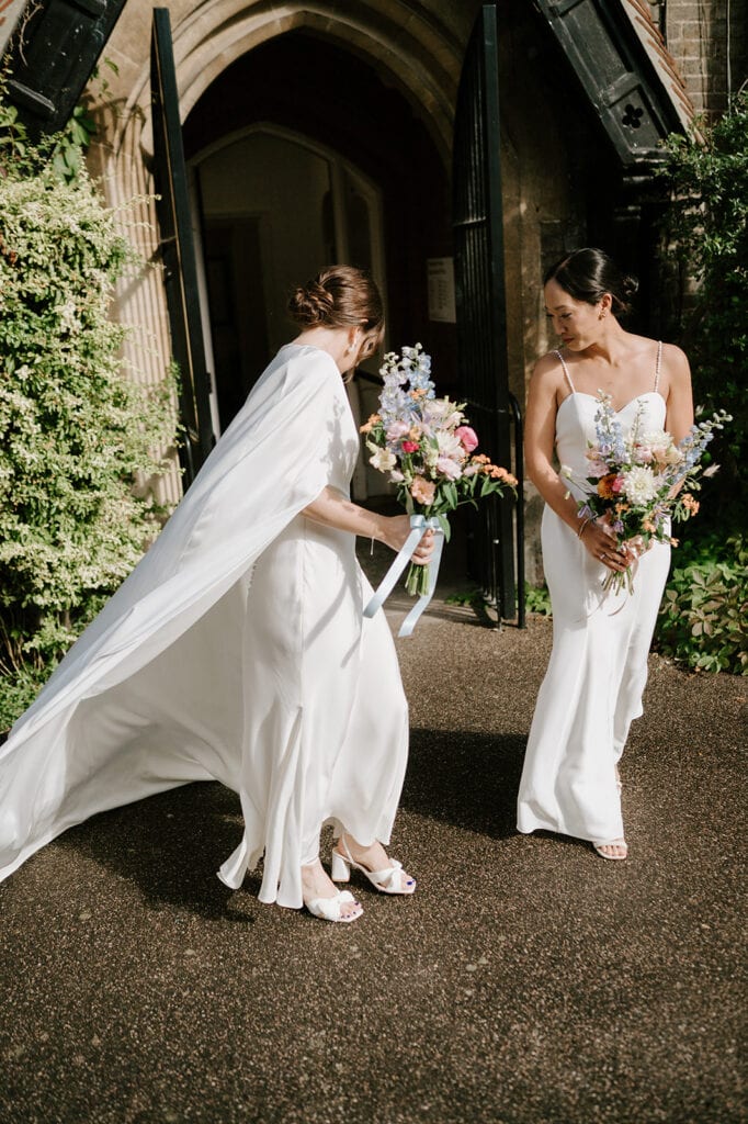 Two women in elegant white wedding dresses stand outside a building with large wooden doors in Broadstairs. Each holds a colorful bouquet of flowers, smiling amidst the greenery, as sunlight casts gentle shadows on the ground. Image by Pearce Wedding Photography.