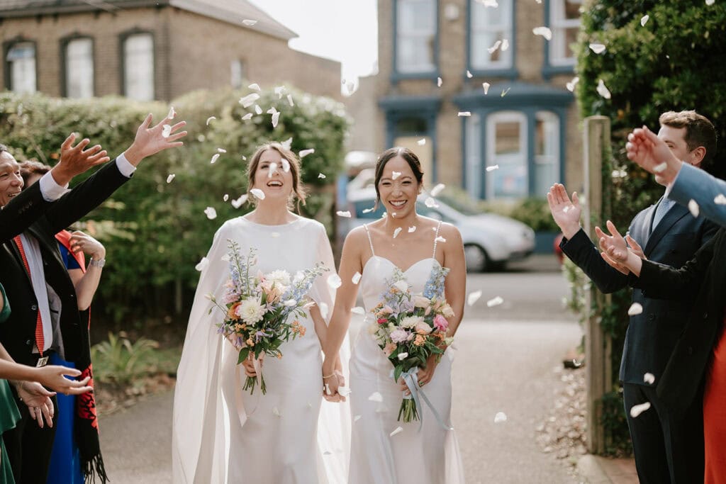Two brides in white dresses hold hands and smile joyfully as guests throw confetti at their Broadstairs wedding. Each holds a bouquet of colorful flowers. The scene is outdoors, with cheering friends celebrating around them against a backdrop of buildings and greenery. Image by Pearce Wedding Photography.