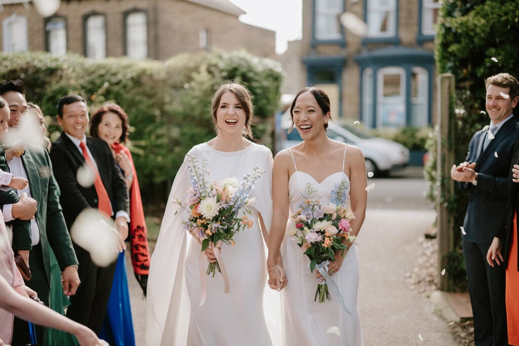 Two brides in white dresses holding bouquets walk joyfully between guests throwing flower petals at their Broadstairs wedding. They're surrounded by well-dressed friends and family, all smiling and celebrating in a sunny outdoor setting with buildings in the background. Image by Pearce Wedding Photography.