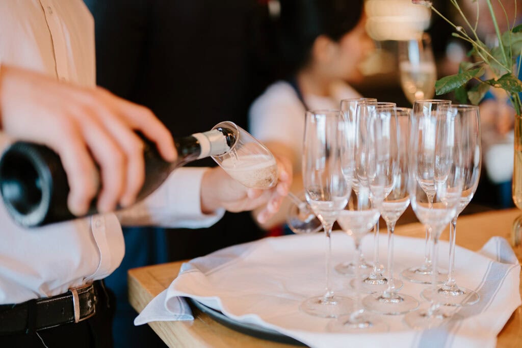 At a Broadstairs wedding, a person in a white shirt elegantly pours champagne into a glass from a bottle. Several empty glasses await on a tray covered with a white cloth. In the background, a blurred figure and part of a floral arrangement add to the celebratory atmosphere. Image by Pearce Wedding Photography.