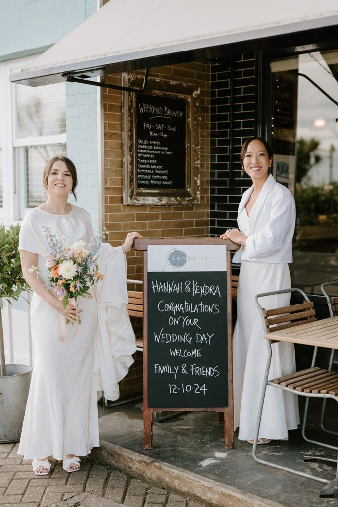 Two people in white dresses stand outside a café in Broadstairs, next to a chalkboard sign. One holds a bouquet, and the sign reads, "Hannah & Kendra, Congratulations on your wedding day. Welcome family & friends. 12-10-24." Tables and chairs are visible outside. Image by Pearce Wedding Photography.