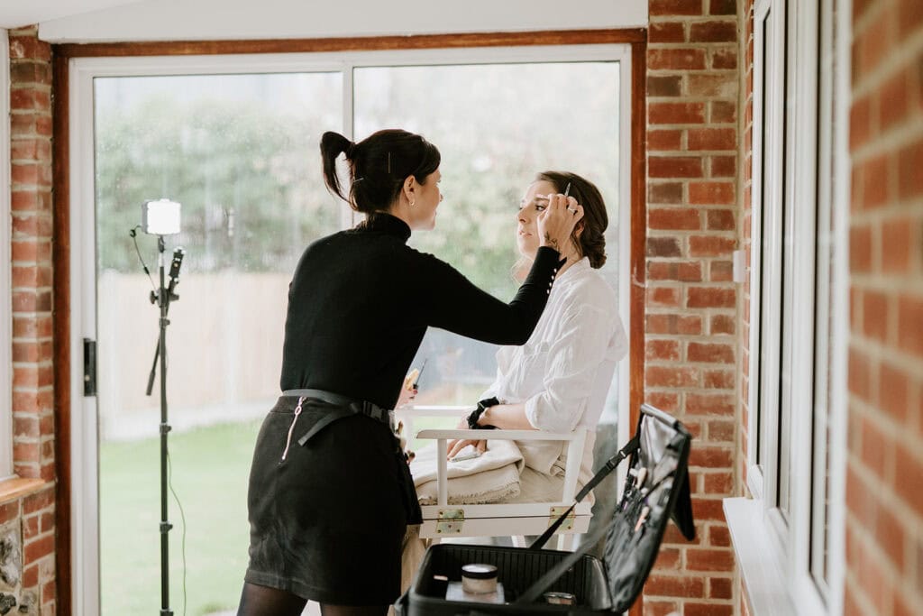 Two women apply makeup using brushes in front of a large window. One woman is seated while the other stands, preparing for a wedding. The setting is a bright room with brick walls and greenery visible outside, possibly in Broadstairs. A makeup case and lighting equipment are nearby. Image by Pearce Wedding Photography.