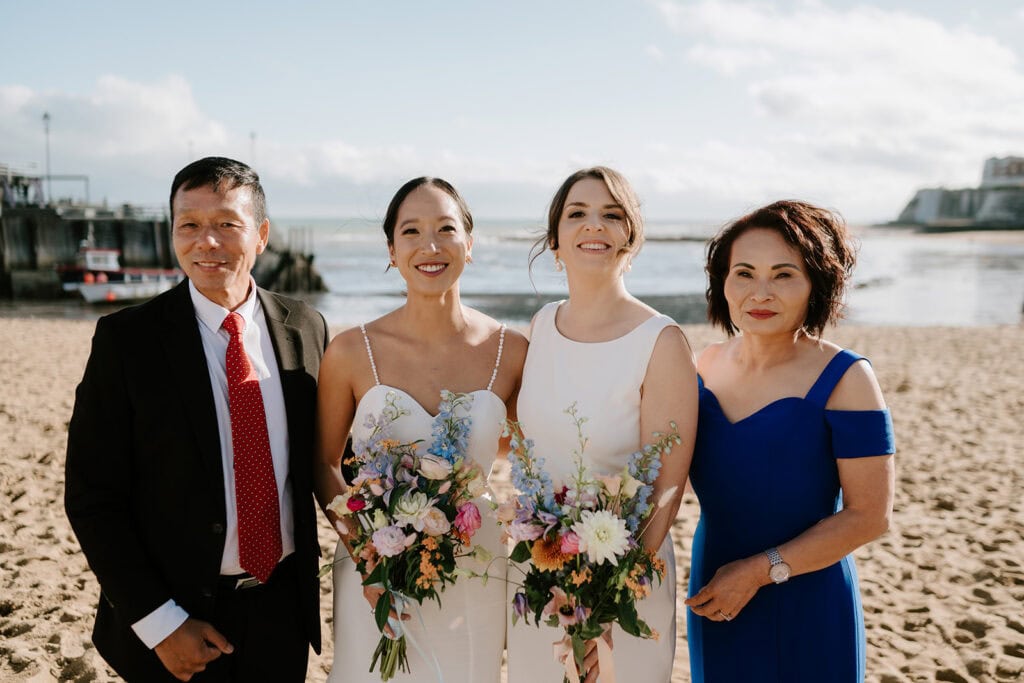 Two brides in wedding dresses holding bouquets stand on a sandy beach in Broadstairs, flanked by a man in a suit and a woman in a blue dress. The background features the ocean and a clear sky on this perfect wedding day. Image by Pearce Wedding Photography.