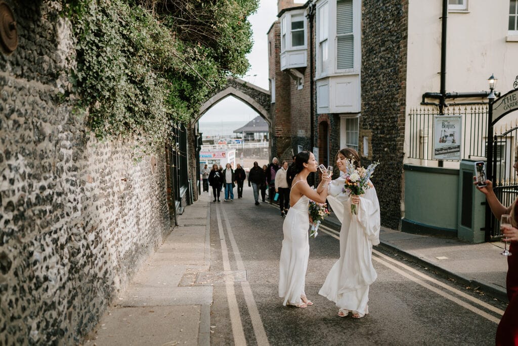 Two people in white dresses share a joyful wedding moment on a narrow Broadstairs street. They are surrounded by stone buildings and a distant archway. A small group of people walk in the background, adding to the lively atmosphere. Image by Pearce Wedding Photography.