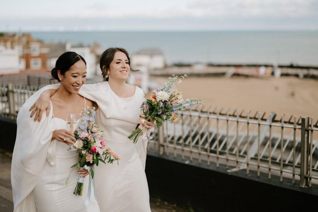 Two women in white dresses, smiling and walking arm in arm, hold floral bouquets and glasses. They appear in a seaside wedding setting at Broadstairs, with a beach, water, and buildings in the background under a partly cloudy sky. Image by Pearce Wedding Photography.