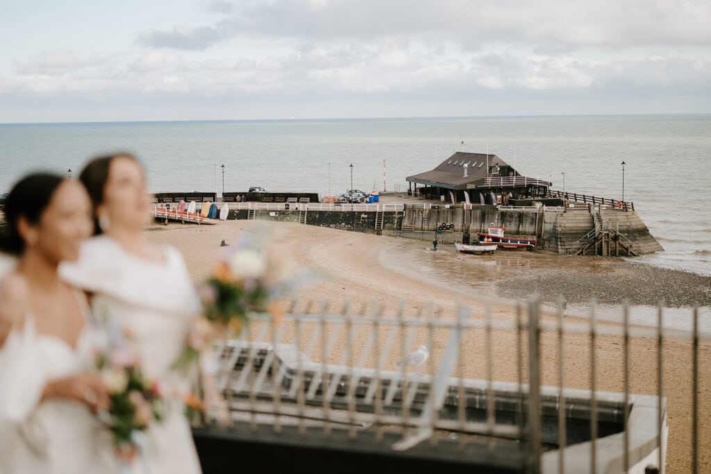 Two people in wedding attire stand with blurred focus in the foreground, holding bouquets. In the background, a coastal view of Broadstairs features a sandy beach, a pier with a small building, and a calm sea under a cloudy sky. Image by Pearce Wedding Photography.