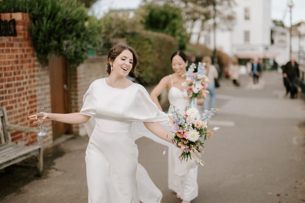 Two joyful brides in white dresses stroll outside a charming Broadstairs venue. The front bride holds a champagne flute, and both have floral bouquets. They are smiling against the backdrop of a brick wall, with wedding guests visible in the distance on the street. Image by Pearce Wedding Photography.