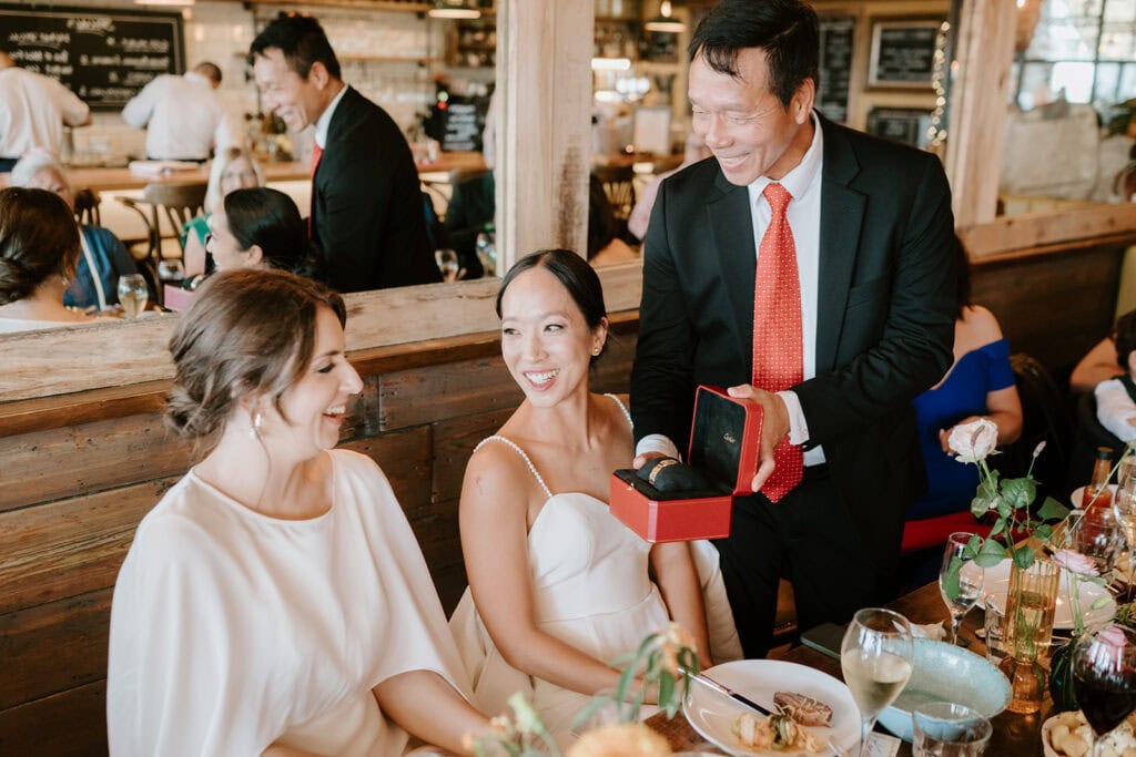 A joyful scene at a lively Broadstairs restaurant: A smiling man in a suit presents a small gift box to a seated woman in a white dress, their faces glowing like it's their wedding day. Another woman beside her looks on happily. The room buzzes with activity, and tables are adorned with plates and glasses. Image by Pearce Wedding Photography.