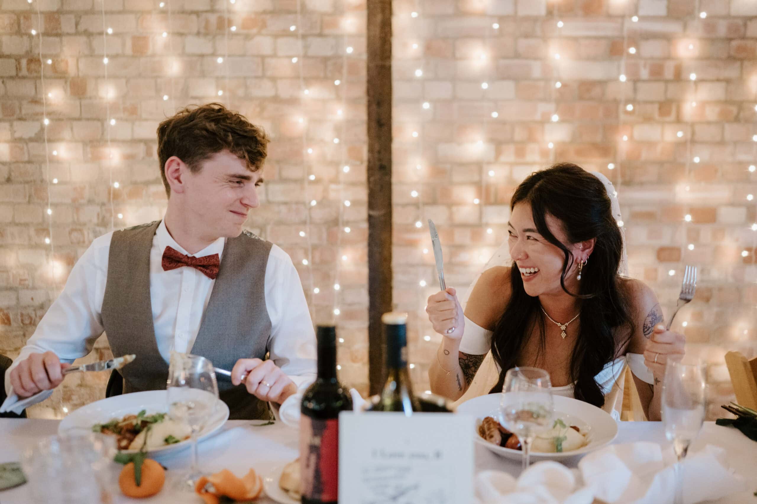 A joyful couple sits at a table during a festive meal, their smiles captured perfectly as if by a Kent wedding photographer. Holding utensils, they are surrounded by plates of delicious food. The brick wall and string lights create a warm, inviting backdrop for this special moment. Image by Pearce Wedding Photography.